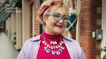 A woman in a bright pink top and lilac jacket with blue glasses with a pink and blue necklace that says 'I'm registered to vote' she is front of a brick building